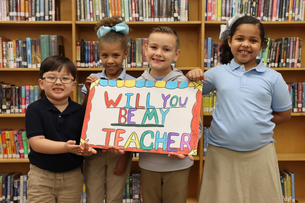 Children hold a sign that says "Will you be my teacher?"